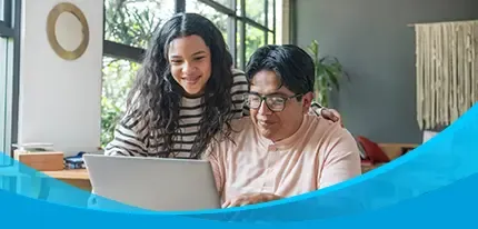 A man and teenage girl smile together while using a laptop at a home dining table.